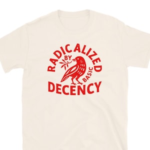 May include: Cream-colored t-shirt with a red graphic. The graphic features a bird holding a sprig, surrounded by the words "RADICALIZED BY BASIC DECENCY" in a circular arrangement. The shirt has short sleeves and a crew neck.