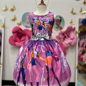 May include: A sleeveless purple and pink dress with a silver bow belt and a matching tiara. The dress features a graphic of animated characters. The dress is displayed on a mannequin.