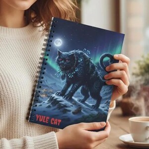 May include: A spiral-bound notebook featuring a dark, mystical illustration of a black cat standing on a snowy mountain under a full moon and aurora borealis, with the text YULE CAT printed in red at the bottom of the front cover.