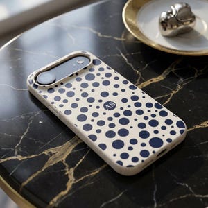 May include: A phone case with a cream-colored background and a pattern of navy blue polka dots. The case has a camera cutout and a circular monogram with the letters "AS". The case is on a black marble surface.