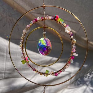 May include: A decorative suncatcher with three gold-colored metal rings of varying sizes. The rings are adorned with colorful beads, crystals, and small flowers. A large, iridescent crystal hangs from the center, reflecting light. A delicate, handcrafted piece.