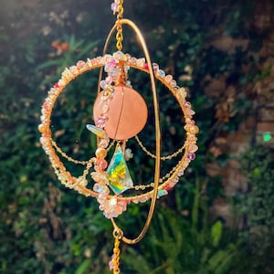 May include: A handcrafted, celestial-themed mobile featuring a large, round, pink rose quartz crystal at its center. The mobile is adorned with gold-colored wire, colorful beads, and iridescent accents, creating a whimsical and decorative piece.