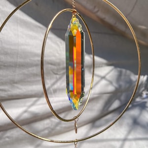 May include: A hanging suncatcher with a large, multi-faceted crystal pendant. The crystal is a prism of colors, including orange, yellow, and blue. It hangs from a gold-colored metal ring and chain. Additional small beads and charms are attached.
