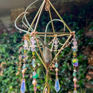 May include: A geometric gold-colored metal air plant holder with a pink flower and dangling iridescent crystals and beads. The air plant is nestled inside the geometric frame, with long green leaves extending outward. The hanging decor is designed for indoor or outdoor use.