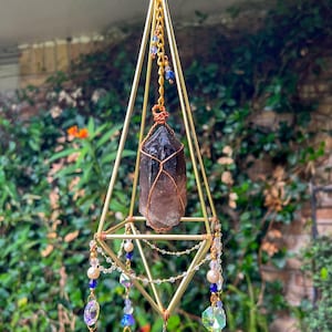 May include: A geometric gold-colored metal prism with a dark brown crystal suspended inside. The prism is adorned with blue, clear, and iridescent crystal beads, pearls, and a pink flower at the top. The crystal is wrapped in copper wire.