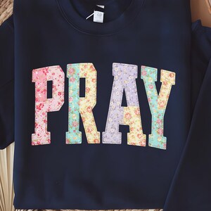 May include: A navy blue sweatshirt featuring the word "PRAY" in large, floral-patterned letters. The letters are in a collegiate style, with a mix of pink, yellow, and purple floral designs. The sweatshirt is displayed on a woven surface.