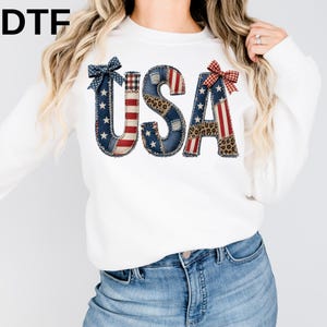 May include: White sweatshirt with the word "USA" in a patchwork design. The letters feature patterns of the American flag, denim, and leopard print, with decorative bows. The sweatshirt is a casual, patriotic apparel item.