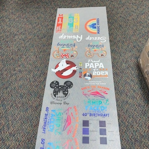 Peut inclure: Une feuille d'autocollants imprim&eacute;s avec divers motifs et textes. Les motifs incluent un logo Ghostbusters, des oreilles de Mickey Mouse et des phrases comme "Proud Papa of 2023" et "Ship Faced !" en plusieurs couleurs. La feuille comprend &eacute;galement des &eacute;chantillons de couleurs.