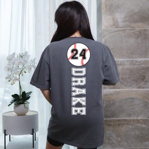 May include: A dark gray t-shirt with a white baseball graphic and the number 24. The name "DRAKE" is printed vertically down the back of the shirt.