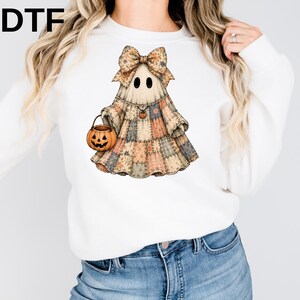 May include: A white long-sleeve sweatshirt featuring a graphic of a ghost wearing a patchwork dress and a large bow. The ghost holds a small orange jack-o'-lantern bucket and wears a tiny pumpkin necklace. The text DTF is printed in the top left corner.