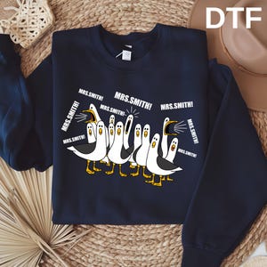 May include: A navy blue crewneck sweatshirt featuring a graphic of multiple white seagulls with yellow beaks and feet. The birds are depicted with open mouths, and the text "MRS.SMITH!" is repeated multiple times around them in white font.