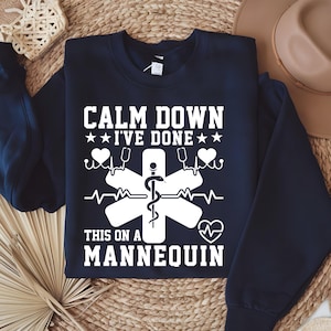 May include: A navy blue crewneck sweatshirt featuring white screen-printed text that reads "CALM DOWN I'VE DONE THIS ON A MANNEQUIN" along with a graphic of a Star of Life symbol, a snake-entwined staff, heart shapes, and an EKG heartbeat line.