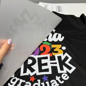K&ouml;nnte beinhalten: Ein schwarzes T-Shirt mit einem wei&szlig;en Design, auf dem "Pre-K Graduate 2023" mit einer regenbogenfarbenen Abschluss-Kappe und Zahlen steht.
