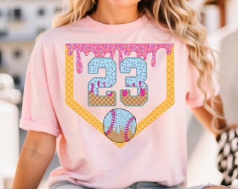 May include: A pale pink t-shirt with a baseball-themed graphic. The design features the number 23 in blue with pink sprinkles, a baseball, and a home plate shape with a waffle cone texture.