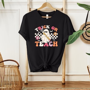 Peut inclure: T-shirt noir avec un motif sur le th&egrave;me d'Halloween. Le motif repr&eacute;sente un fant&ocirc;me tenant un crayon, avec les mots "Trick or Teach" dans une police r&eacute;tro. Le motif comprend des fleurs et un motif &agrave; carreaux.