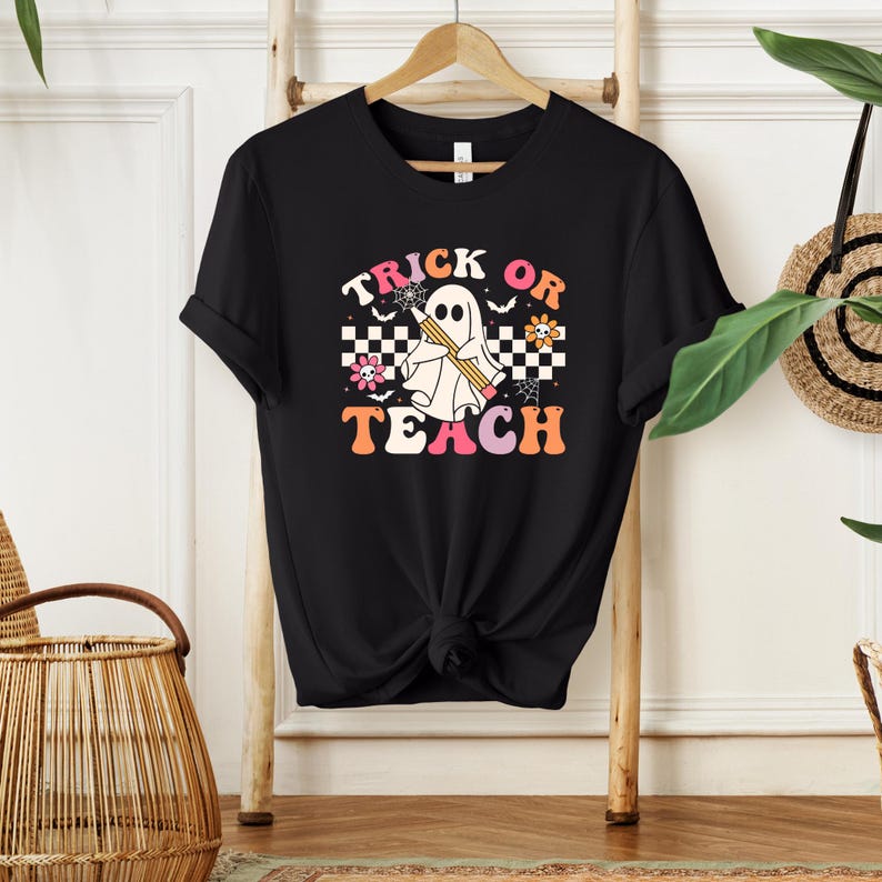Peut inclure: T-shirt noir avec un motif sur le th&egrave;me d'Halloween. Le motif repr&eacute;sente un fant&ocirc;me tenant un crayon, avec les mots "Trick or Teach" dans une police r&eacute;tro. Le motif comprend des fleurs et un motif &agrave; carreaux.