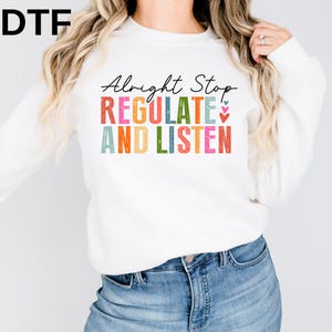 May include: A white long-sleeve crewneck sweatshirt featuring the black cursive text Alright Stop and the colorful block letters REGULATE AND LISTEN with a distressed texture. Three small stacked hearts in blue, pink, and red appear on the right.