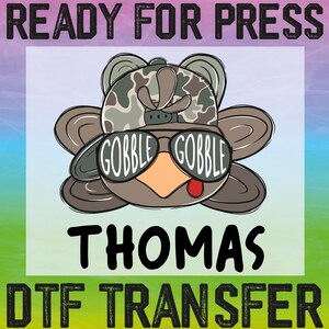 May include: A cartoon turkey wearing sunglasses that say "GOBBLE GOBBLE" and a camouflage hat. The turkey is named "THOMAS" and the image says "READY FOR PRESS" and "DTF TRANSFER". The turkey has a red tongue sticking out.