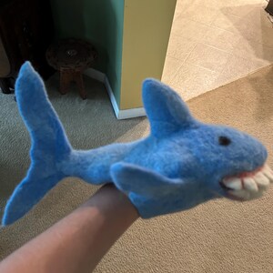 May include: A blue felt shark puppet with a wide open mouth revealing white teeth and pink gums. The puppet has a dorsal fin, tail fin, and side fins. The shark puppet is designed to fit over a hand.