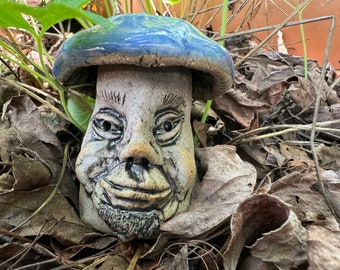 Puede incluir: Una figurita de hongo de cerámica con un sombrero esmaltado azul y blanco y una cara detallada y caprichosa. El hongo está sentado en un lecho de hojas marrones.