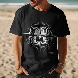 Pode incluir: Camiseta preta com um gráfico branco de um avião na frente de um design de sol radiante. O texto "Proudly Serving" é impresso abaixo do gráfico.