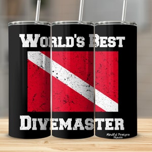 Puede incluir: Un vaso de acero inoxidable negro con un gráfico blanco que dice "World's Best Divemaster" y una bandera de buceo roja y blanca.