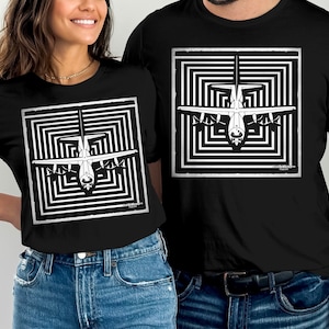 May include: Black t-shirts featuring a white graphic of an airplane within a square, surrounded by a black and white geometric pattern. The design is centered on the front of the shirts. The shirts are worn by two people.