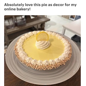 Könnte beinhalten: Ein Zitronenkuchen mit gelber Füllung, garniert mit Schlagsahne und einer Zitronenscheibe. Der Kuchen hat einen weißen Schlagsahnerand und einen Kruste mit einem braunen, strukturierten Rand. Der Text lautet: "Ich liebe diesen Kuchen als Dekoration für meine Online-Bäckerei!"