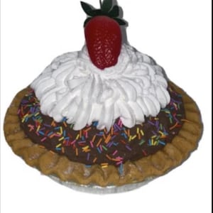 Puede incluir: Una tarta decorativa con relleno de chocolate, cubierta con glaseado blanco, chispas de colores y una fresa roja. La tarta tiene una corteza dorada y se presenta sobre una superficie plateada. Un dulce.