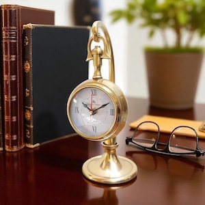 Puede incluir: Un reloj de escritorio dorado con esfera blanca y números romanos negros. El reloj tiene una esfera redonda y un asa curva. El reloj está sobre una base dorada. El reloj está junto a un par de gafas y algunos libros.