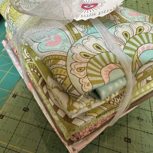 May include: A bundle of patterned fabric squares, tied with a silver ribbon. The fabric displays a floral design in green, pink, and blue hues. A tag with an eye design and the text "tula pink" is attached to the ribbon.