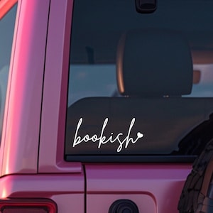 May include: A white vinyl decal applied to the rear window of a pink vehicle. The decal features the word bookish written in a cursive script font, with a small heart symbol positioned at the end of the text, creating a simple graphic design.