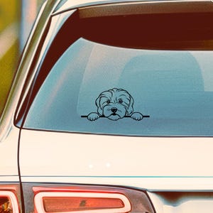 May include: A black vinyl car decal featuring a stylized illustration of a dog peeking over a horizontal line. The dog has a fluffy appearance with detailed facial features. The decal is applied to the rear window of a white car.