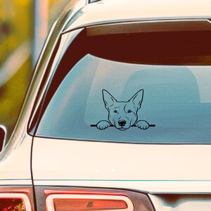 May include: Black vinyl car decal featuring a dog's head peeking over a horizontal line. The dog has pointed ears and a detailed face. The decal is applied to the rear window of a white car.