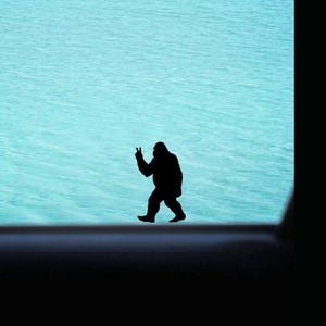 May include: A black silhouette of a Bigfoot figure giving a peace sign, set against a backdrop of turquoise water. The figure is walking, with the water visible through a window frame.