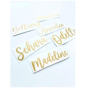May include: White rectangular stickers with gold cursive lettering. The stickers display names including Jennifer, Bellamy, Annalee, Sehara, Odella, and Madeline. The stickers are arranged on a white surface.