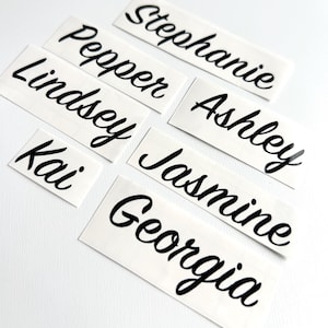 May include: A collection of six individual black vinyl adhesive decals featuring names written in a cursive script font. The names displayed on the rectangular white backing paper are Stephanie, Pepper, Ashley, Lindsey, Jasmine, and Kai, with Georgia positioned at the bottom.