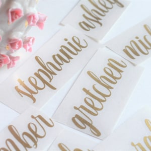 May include: Gold calligraphy name labels on white paper. The names are Stephanie, Ashley, Gretchen, Michelle, Amber, and Sarah.