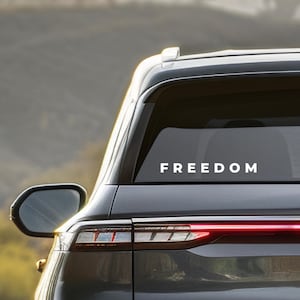 May include: A car's rear window displays the word "FREEDOM" in white, bold lettering. The vehicle is dark gray, with a side mirror visible. The background is blurred, suggesting an outdoor setting.