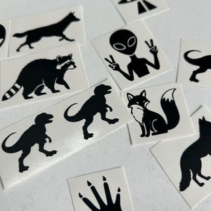 May include: A collection of black and white stickers featuring various designs. Included are silhouettes of a wolf, raccoon, fox, dinosaurs, an alien giving a peace sign, a UFO, and a paw print.