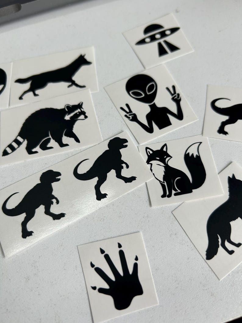 May include: A collection of black and white stickers featuring various designs. Included are silhouettes of a wolf, raccoon, fox, dinosaurs, an alien giving a peace sign, a UFO, and a paw print.