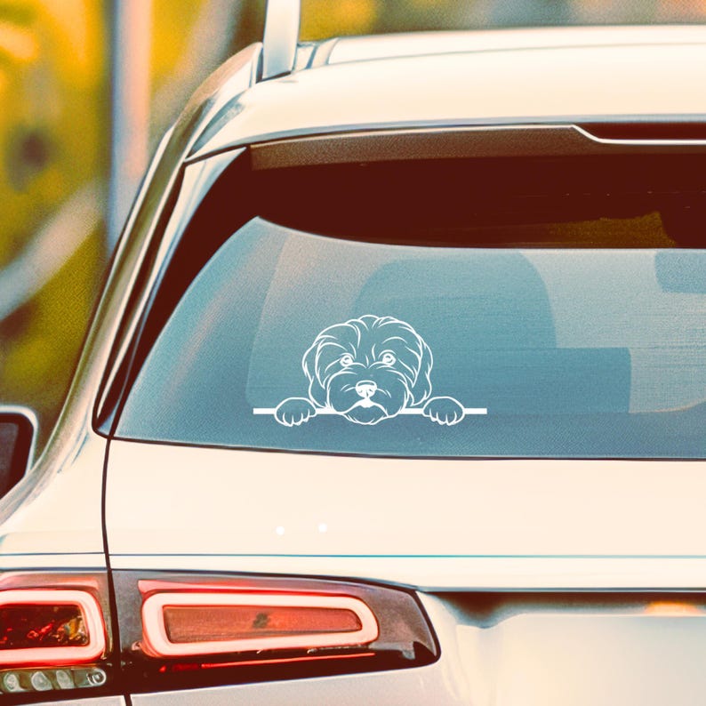 K&ouml;nnte beinhalten: Wei&szlig;er Vinyl-Autoaufkleber mit einer stilisierten Illustration eines flauschigen Hundes, der &uuml;ber eine horizontale Linie schaut. Das Gesicht und die Pfoten des Hundes sind sichtbar, was ein verspieltes Design ergibt. Der Aufkleber ist auf einer Autoscheibe angebracht.
