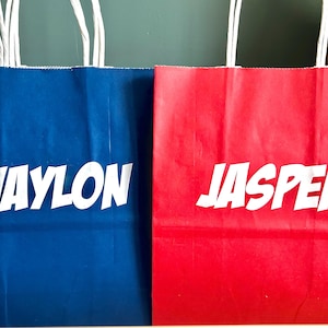 May include: Two paper gift bags, one blue and one red, with white text. The blue bag reads "WAYLON" and the red bag reads "JASPER". Both bags have white rope handles.