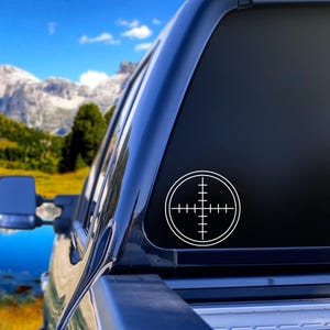 May include: A white vinyl decal of a rifle scope reticle, applied to the rear window of a black truck. The crosshair design is centered within a circle. The background shows a mountain range and blue sky.