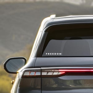 May include: Rear view of a dark gray vehicle with the word "FREEDOM" in white lettering on the rear window. The vehicle's taillight is illuminated, and a side mirror is visible.