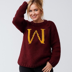 Peut inclure: Pull en tricot bordeaux avec un grand "M" jaune sur le devant. Le pull est porté avec un jean noir et des baskets blanches. La femme sourit et a la main dans les cheveux.