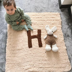 Könnte beinhalten: Eine cremefarbene gestrickte Decke mit einem braunen Filzbuchstaben "H" und einem braunen gehäkelten Hasenspielzeug. Die Decke liegt auf einem grauen Teppich und ein Baby sitzt auf der Decke.
