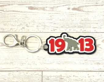 May include: A keychain with a silver clasp and ring. The keychain features a black, red, and gray design with the numbers "1913" and a gray elephant silhouette. The background is a light wood grain.