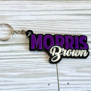 Pode incluir: Chaveiro roxo e prateado com o texto "MORRIS Brown" em um fundo de madeira branco.
