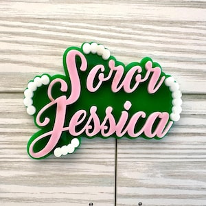 Op de afbeelding: Een gepersonaliseerde acryl taarttopper met de tekst Soror Jessica in lichtroze cursief schrift. De tekst is gelaagd over een donkergroene achtergrond met een geschulpte rand, geaccentueerd door kleine witte ronde kralen langs de omtrek.
